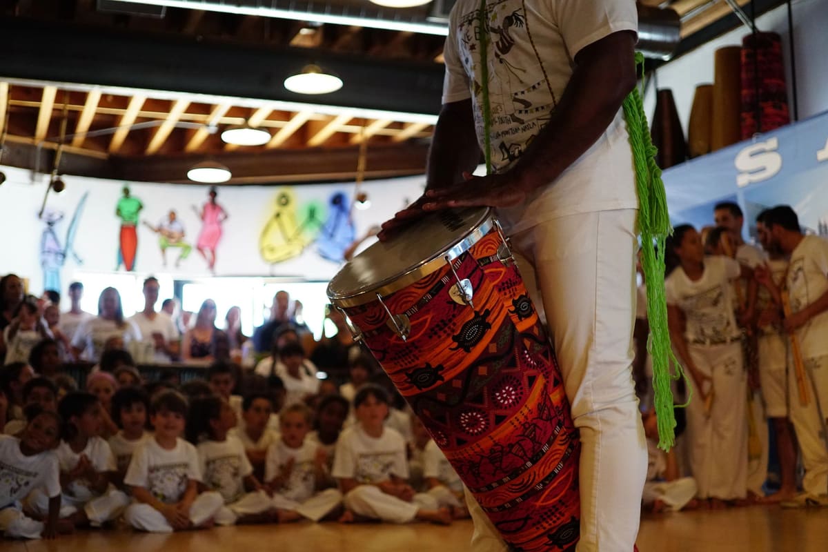 Children in white capoeira uniforms sitting on the floor watching a drummer