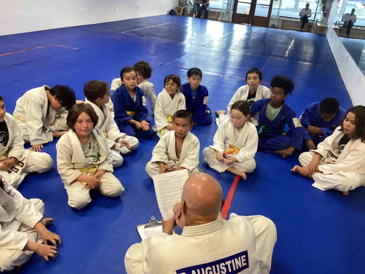 Children in judo uniforms sitting in a circle on the mat listening to their instructor