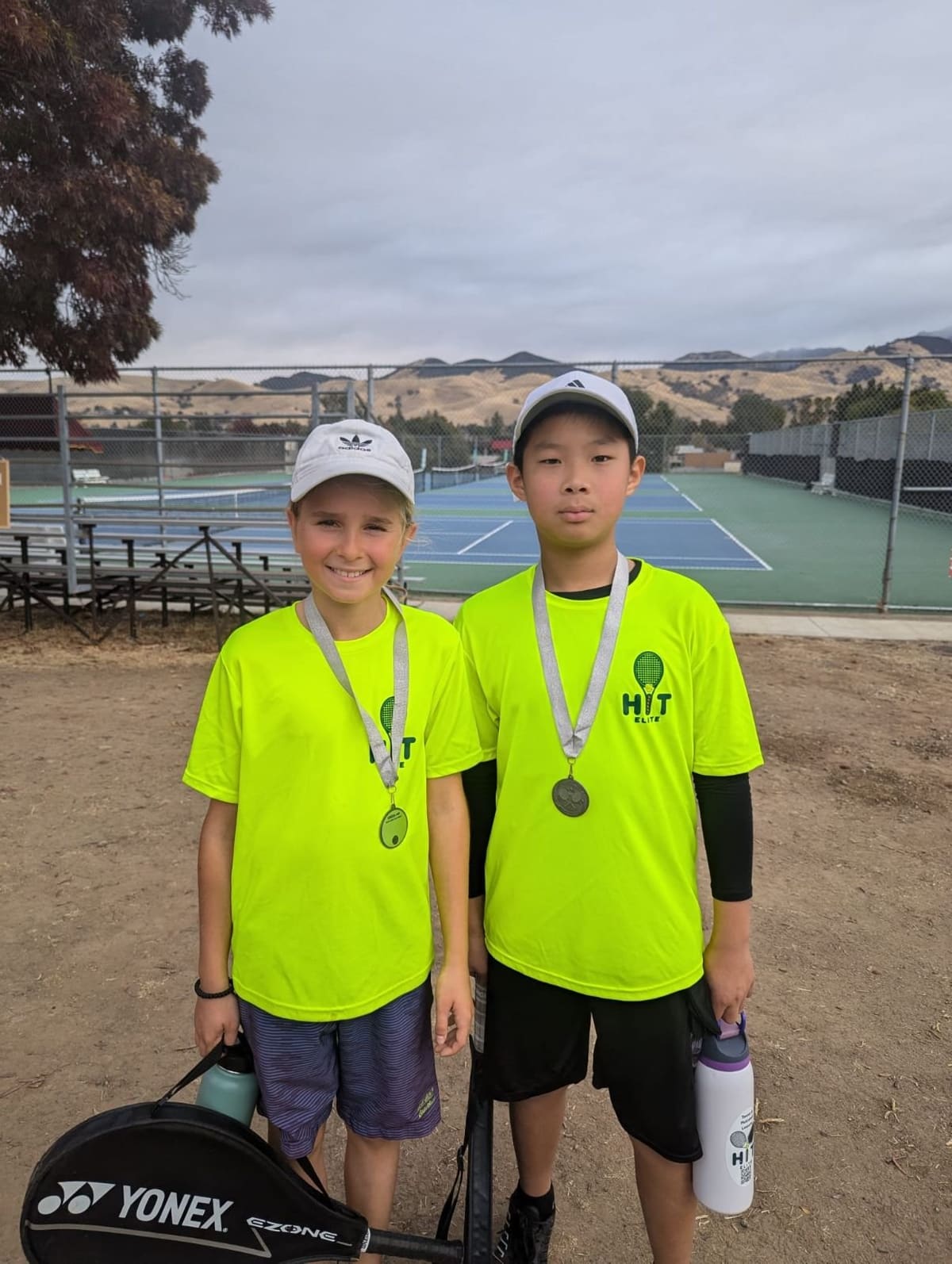 Two young players in matching shirts holding medals on a court
