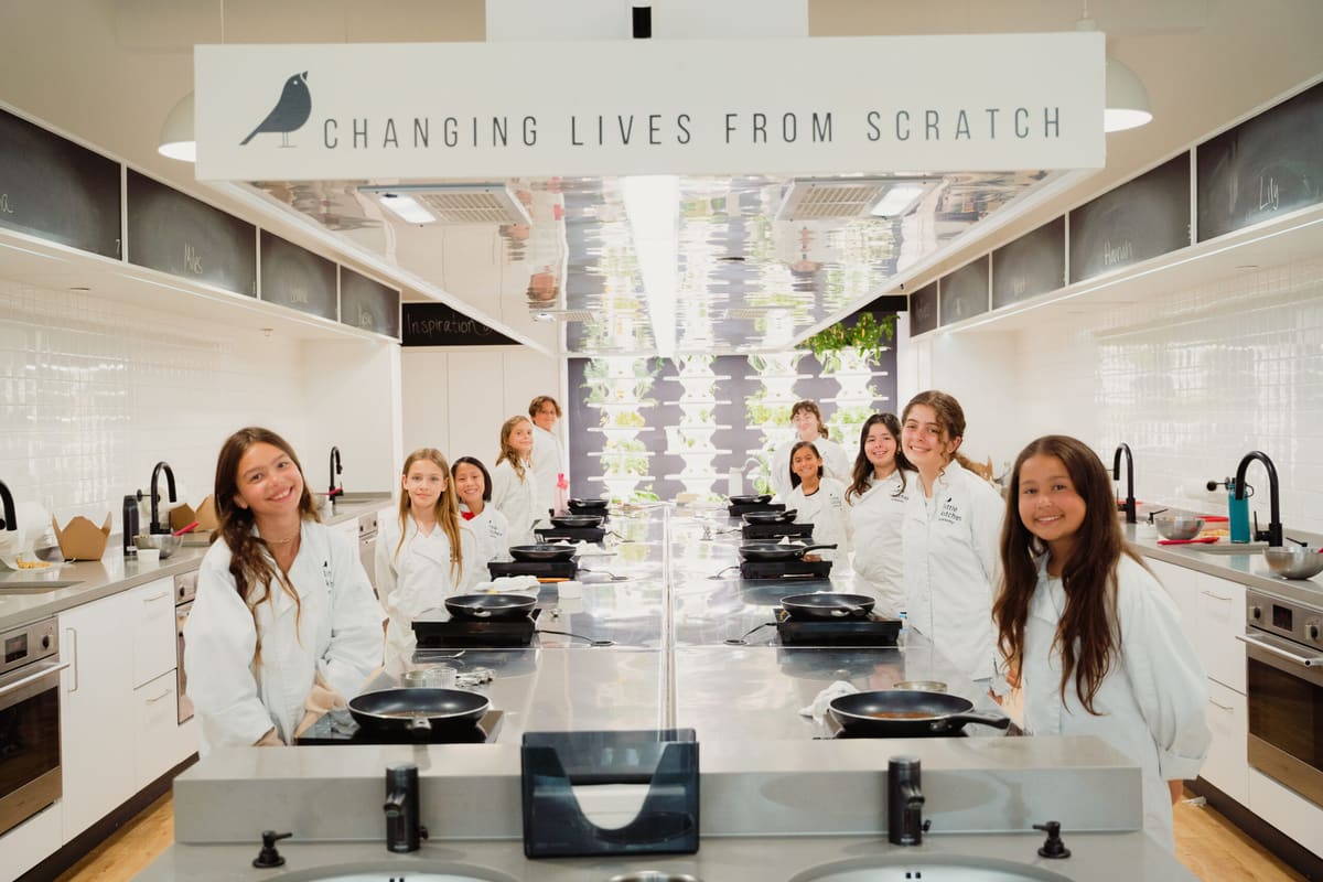 Girls in white chef coats standing at cooking stations in a Little Kitchen Academy kitchen