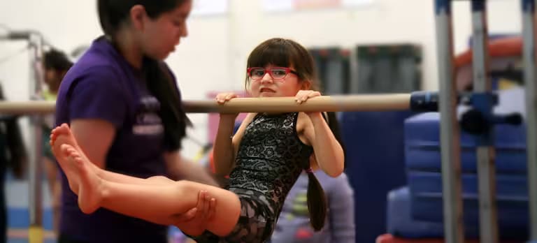 A girl in a leotard hangs from a low bar while a coach supports her legs