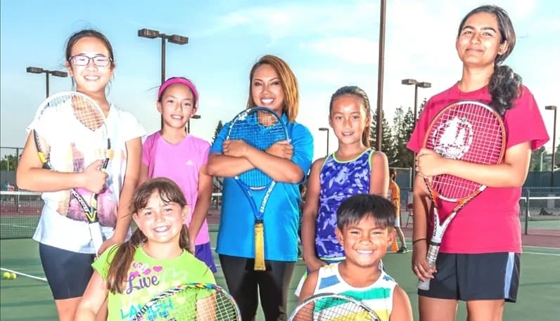 A coach and a group of smiling kids holding tennis racquets on a court