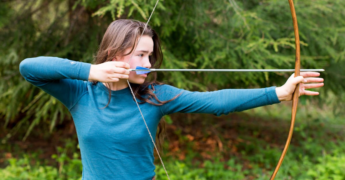 A teenager drawing a wooden bow to shoot an arrow outdoors among trees