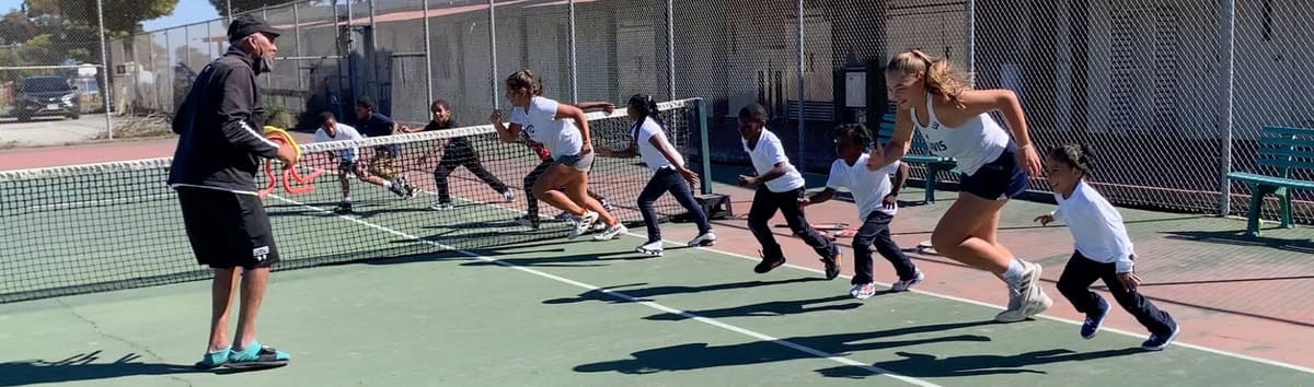 A coach and young players sprint along a net on a court